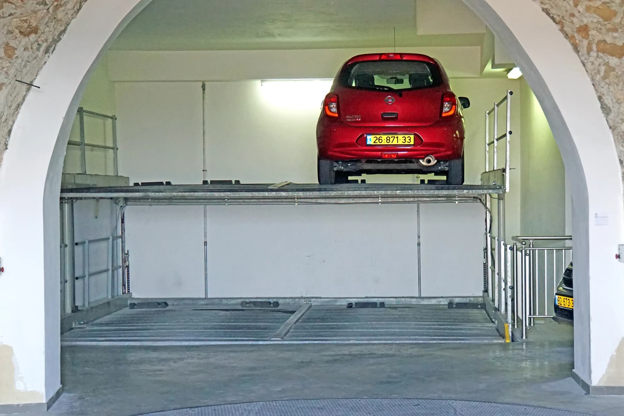 A red car on the raised platform of a two-level pit parking system beneath a stone arch