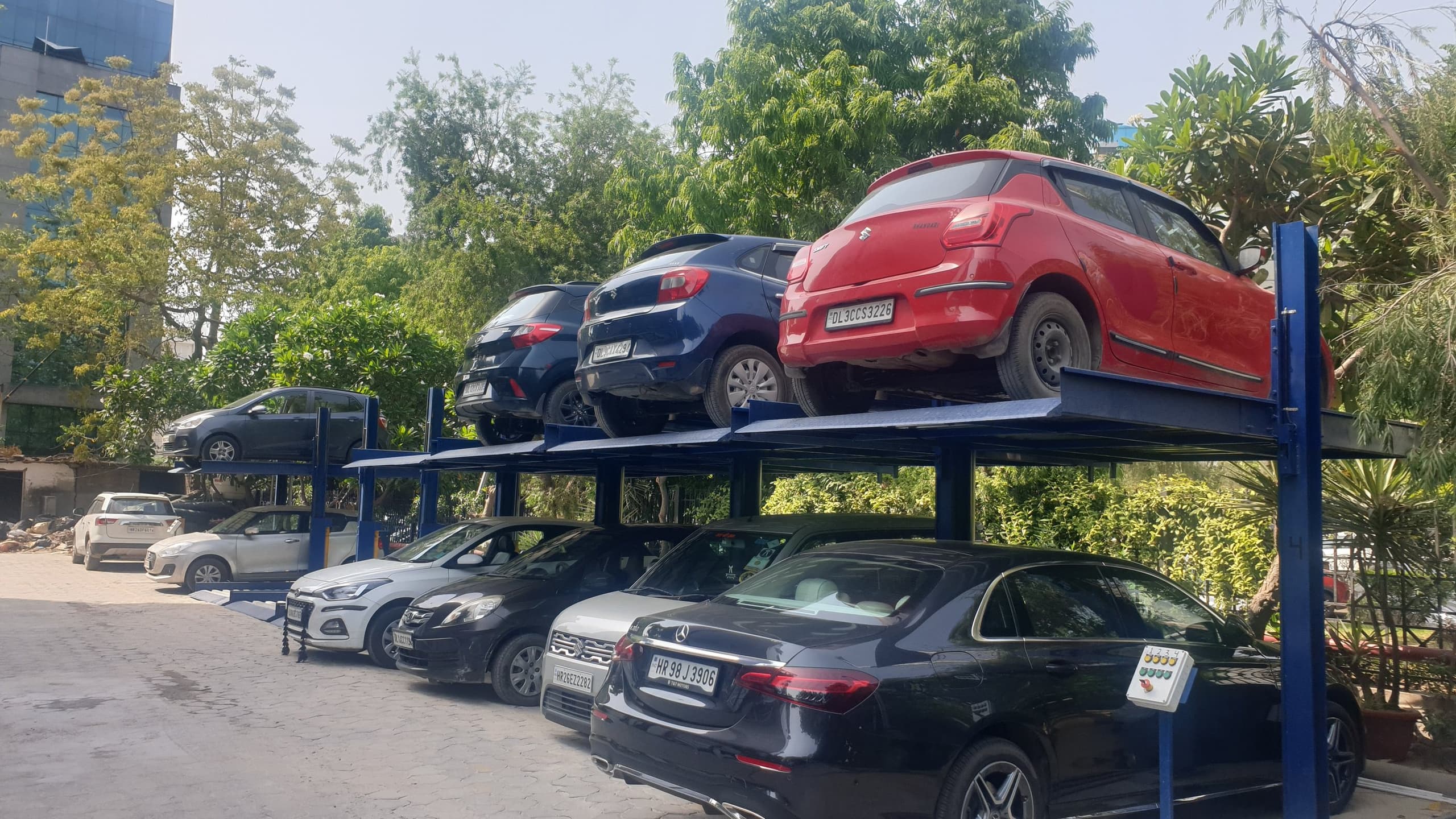 Row of blue two-post hydraulic stack parking units outdoors, upper platforms loaded with cars above cars parked at ground level